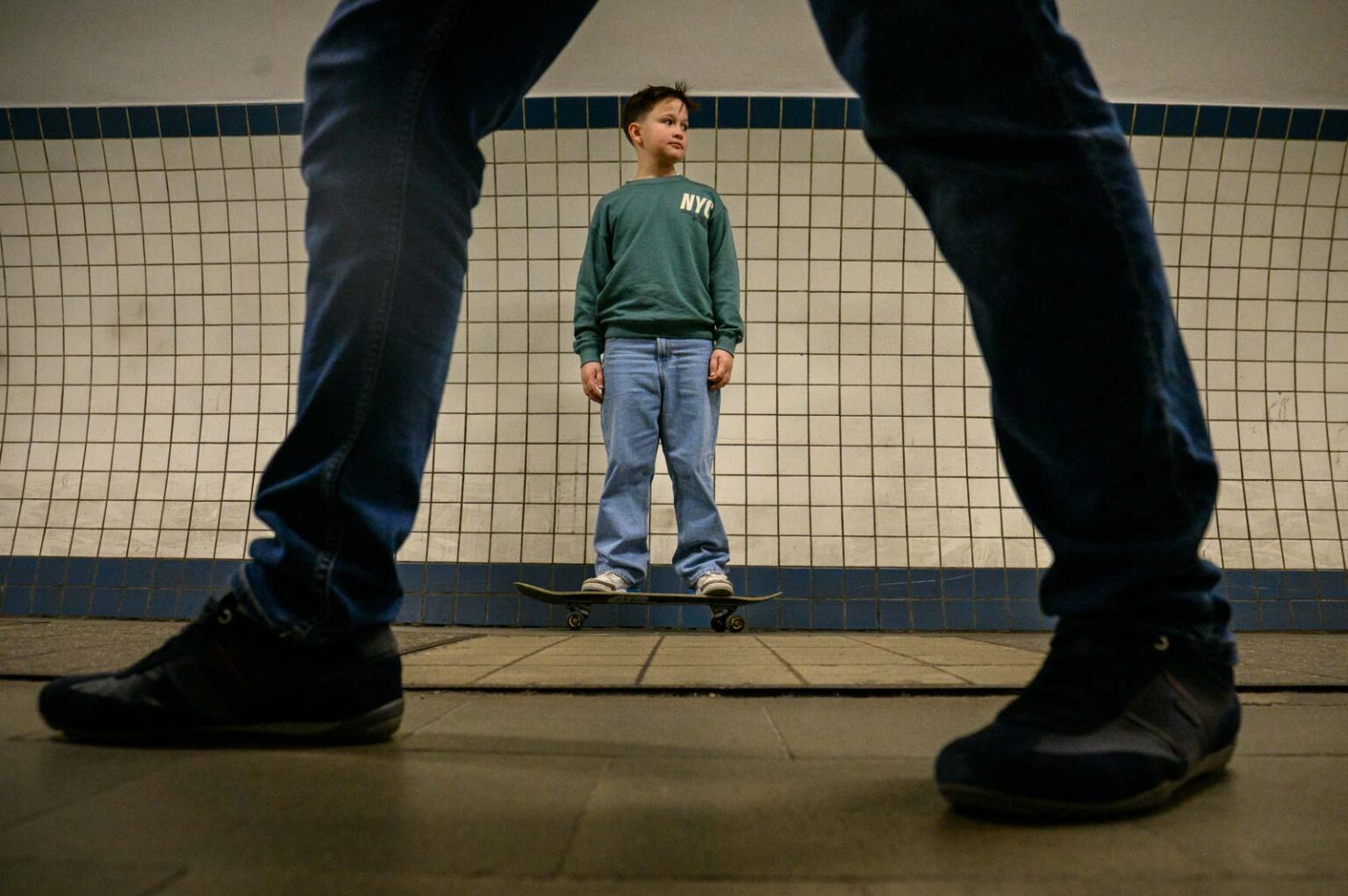 Jongen op skateboard bij betegelde muur, gezien door benen op voorgrond.