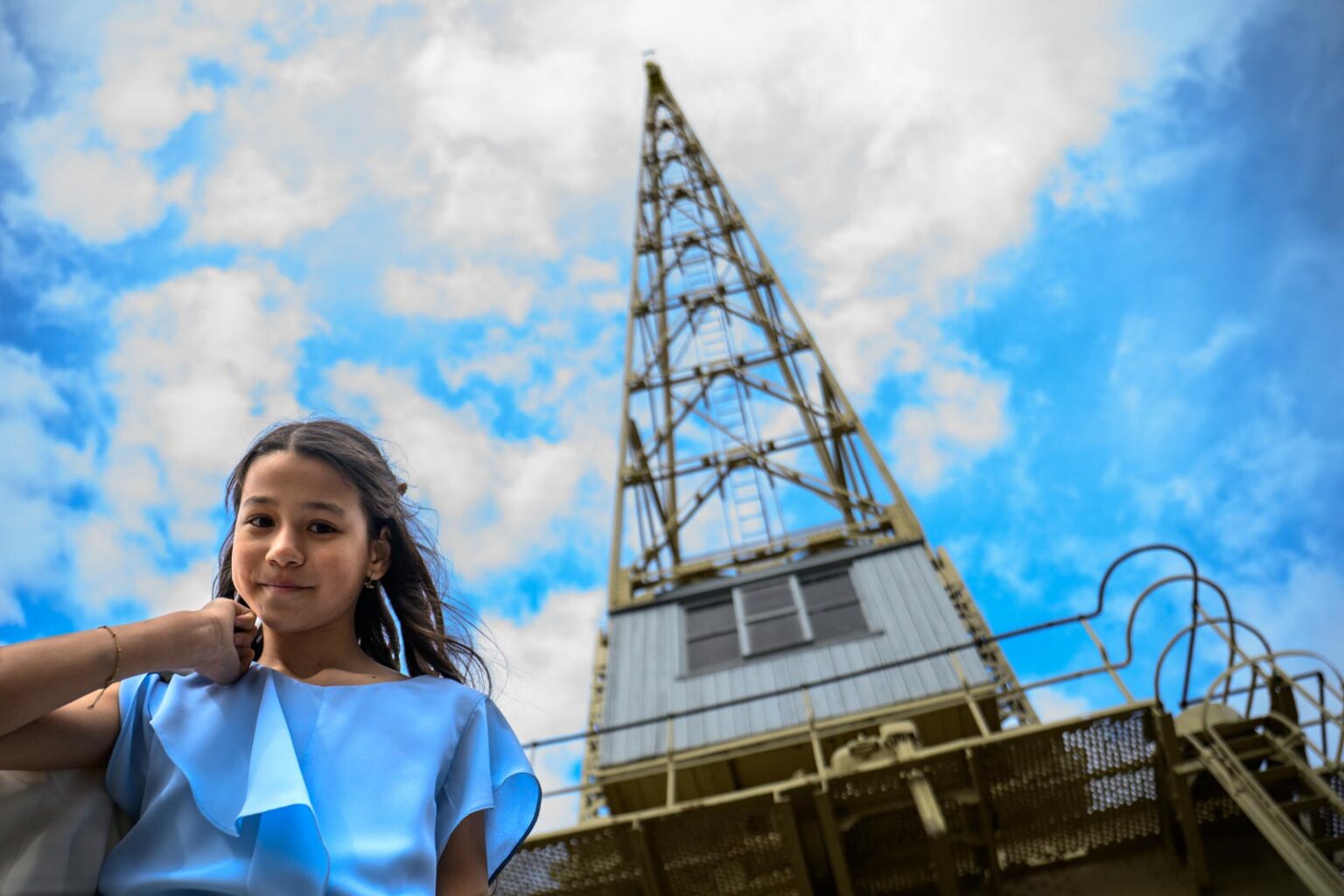 Meisje in blauwe jurk staat voor een hoge metalen toren onder een bewolkte hemel.